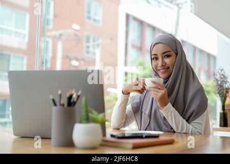 Attractive muslin woman working in office with laptop computer, hand holding cup of coffee. Stock Photo