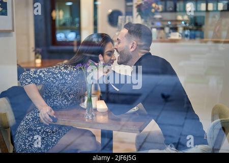 Through glass side view of cheerful couple having fun while sitting at table in modern light cafeteria during romantic date Stock Photo