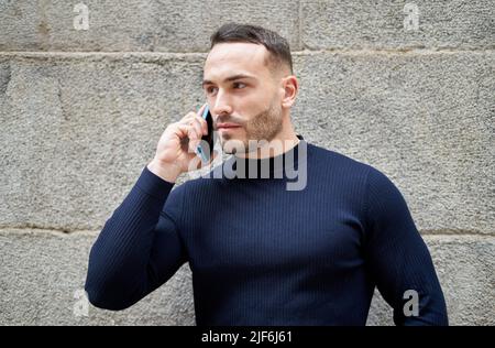 Serious bearded male looking away while standing on wall talking on mobile phone on the street Stock Photo