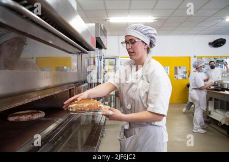 Side view of focused female baker taking out freshly baked bread from hot oven while learning baking in baker school Stock Photo