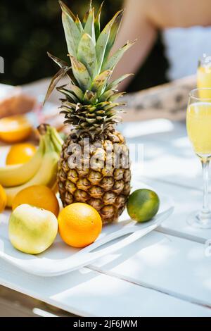 Ripe pineapple with mix of fresh fruits served on white table with beverages on terrace of building on sunny summer day Stock Photo