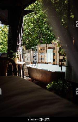 Ceramic bathtub placed near wooden fence on patio of building with bed and roof on sunny summer day in countryside Stock Photo