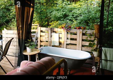 Ceramic bathtub placed near wooden fence on patio of building with bed and roof on sunny summer day in countryside Stock Photo