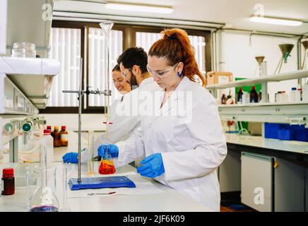 Side view of group of chemists in white uniform and protective glasses preparing chemical liquid while doing research together in modern laboratory Stock Photo
