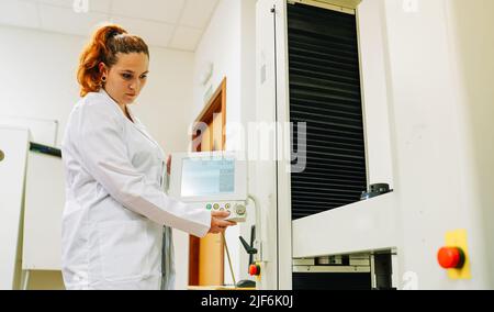 Focused female scientist in medical white uniform turning on modern professional equipment with display while working in modern light lab Stock Photo