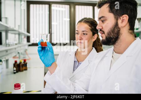Attentive scientists in medical gowns examining beaker with chemical liquid while working together in modern light laboratory with various equipment Stock Photo