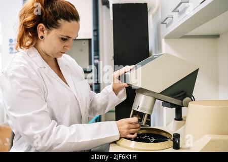 Side view of attentive female worker in medical robe operating modern machine for chemical research while standing in light laboratory Stock Photo