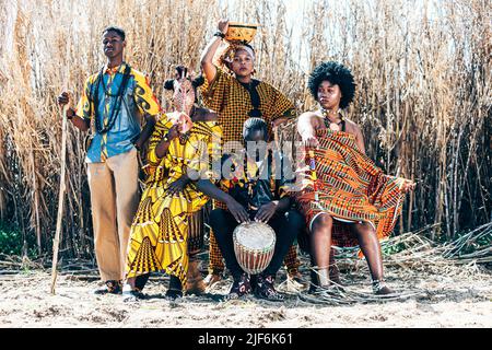 Full body group of black and mixed race women and men in colorful authentic African outfits with sticks and drums sitting and standing against dry fie Stock Photo