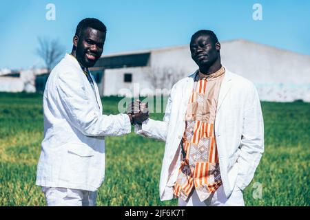 Confident black male models in trendy white suits and traditional ornamental shirts shaking hands and looking at camera while standing in grassy field Stock Photo