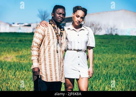Mixed race Woman in casual white dress leaning on shoulder of serious black man in traditional African shirt with necklaces while standing in green gr Stock Photo
