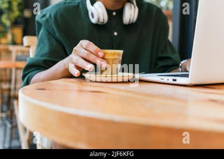 Anonymous ethnic male freelancer drinking coffee while working remotely on modern netbook in cafe Stock Photo