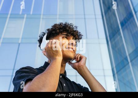 Low angle of young male in modern headphones listening music while standing on city street near tall building Stock Photo