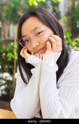 Peaceful young Asian female in stylish eyeglasses looking at camera while sitting on street with green plants on sunny day and leaning on hands Stock Photo