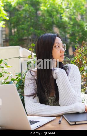 Serious focused Asian female freelancer while sitting at table with netbook and organizer on summer day in street cafe Stock Photo