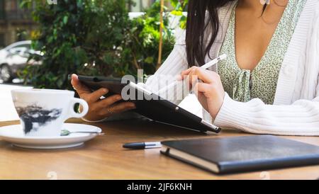 Cropped unrecognizable female freelancer writing on tablet with stylus while sitting on terrace of cafeteria on summer day in city Stock Photo