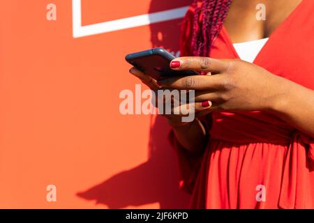 Low angle of crop anonymous African American female in trendy outfit standing on street against red background Stock Photo