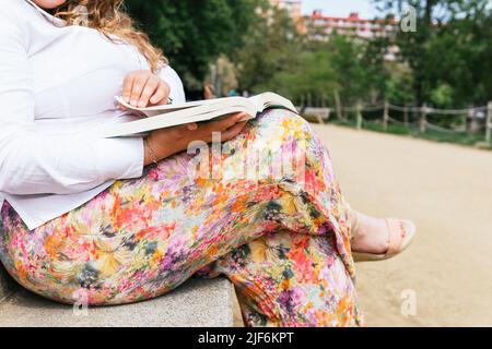 Crop unrecognizable overweight female in colorful pants reading interesting book while sitting on bench in park on summer day in city Stock Photo