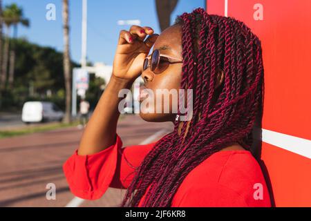Side view of young African American female in trendy outfit and glasses looking at camera while standing against red background Stock Photo