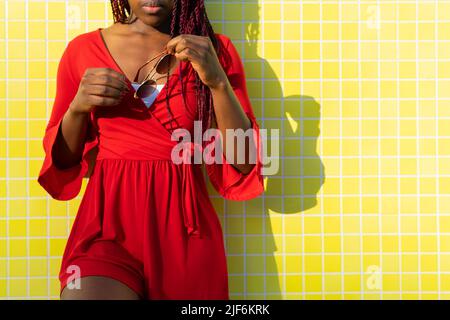 Crop view of anonymous African American young Woman in trendy red dress and braids holding sunglasses against yellow tiled wall Stock Photo