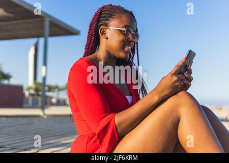 African American young Woman in trendy red dress and braids using phone while sitting at the beach Stock Photo