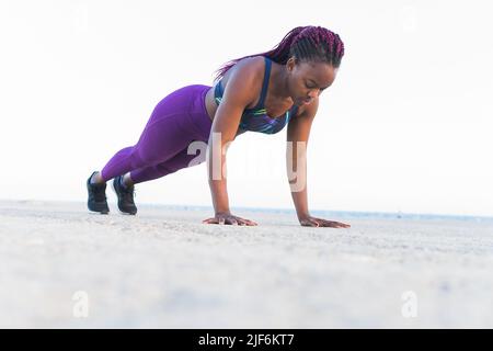 Low angle of full body fit black Woman athlete in sportswear doing push ups exercises at the sea shore Stock Photo