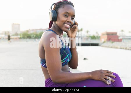Side view of cheerful African American female in sportswear and headphones smiling and looking at camera while sitting on street listening to music Stock Photo