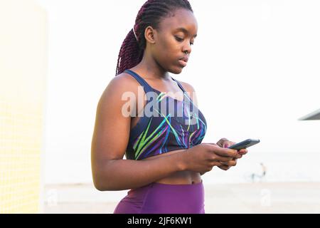 Fit black female athlete in sportswear using phone standing outdoors against neutral background Stock Photo