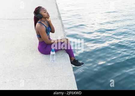 Side view of African American female in sportswear and headphones sitting at pier near sea Stock Photo