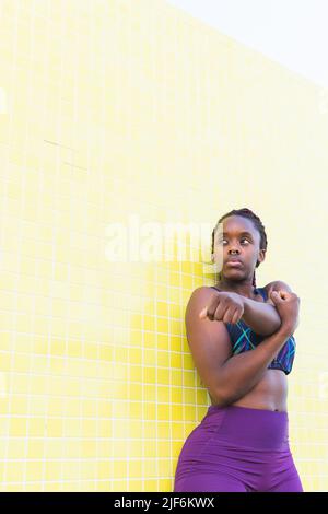 Fit black female athlete in sportswear doing stretching exercises while warming up before training against yellow tiled wall Stock Photo