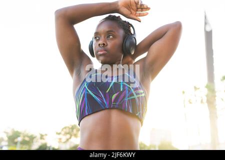 Fit female athlete in sportswear and headphones stretching triceps while warming up before training Stock Photo