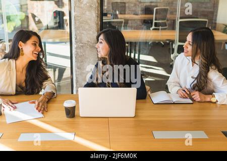 High angle of cheerful Hispanic female entrepreneurs in smart casual clothes smiling and discussing business ideas while sitting at table and working Stock Photo