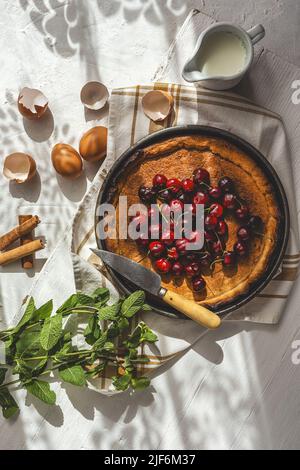 From above baked cherry pie on kitchen cloth near mint leaves, eggs, shells and jug of milk on white background Stock Photo