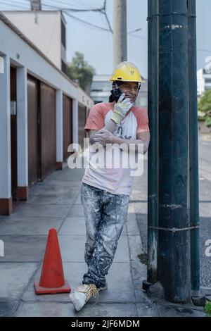 Full length of man in safety hardhat leaning on lamp post and talking on mobile phone on sidewalk Stock Photo