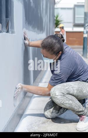 Side view of professional female painter in protective mask in process of plastering wall using special equipment on city street Stock Photo
