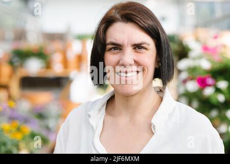 Cheerful adult brunette in white shirt with short hair smiling and looking at camera while standing on blurred background of flower store Stock Photo