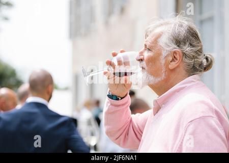 Side view of senior bearded male with gray hair drinking red wine near building during wedding party against people on blurred background Stock Photo