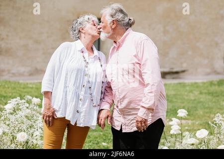 Content elderly couple looking at each other while standing holding hands on street with decoration near building during wedding party on summer day Stock Photo