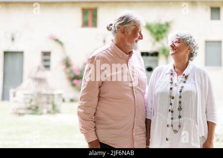 Content elderly couple looking at each other while standing on street with decoration near building during wedding party on summer day Stock Photo