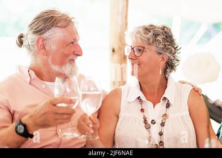 Glad senior couple clinking wineglasses with water and looking at each other while celebrating wedding on terrace against blurred background Stock Photo