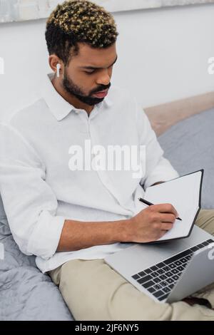 young african american man in wireless earphone making notes near laptop in bedroom Stock Photo