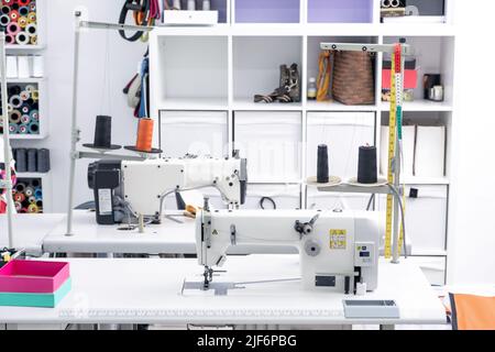 Professional sewing machines with spools of thread placed on white tables in modern light workshop with assorted supplies on shelves in atelier Stock Photo