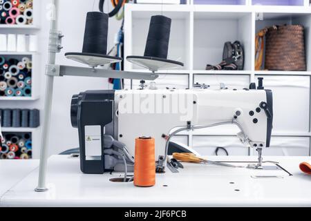 Professional sewing machines with spools of thread placed on white tables in modern light workshop with assorted supplies on shelves in atelier Stock Photo