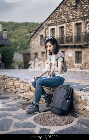 Focused Asian female tourist texting message on cellphone while sitting on stone border near old buildings during trip in Spain Stock Photo