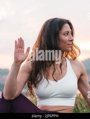 Woman in sportswear sitting on grass and looking away while sitting on green meadow and meditating on sunset Stock Photo