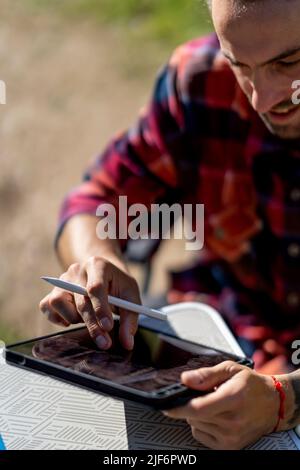 Positive male in casual clothes smiling and browsing data on tablet while sitting at table on sunny summer day in countryside Stock Photo