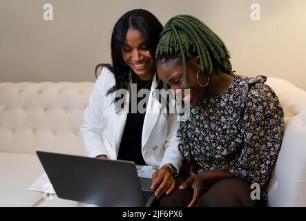 Happy multiracial female entrepreneurs browsing netbook together while working on remote project and sitting on sofa in room Stock Photo