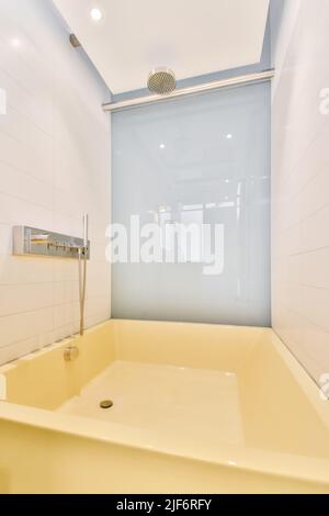 Large square shaped yellow bathtub with chrome shower head in contemporary bathroom with glossy white and blue tiled walls Stock Photo