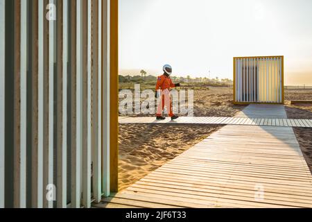 Side view of unrecognizable cosmonaut in spacesuit and helmet walking on boardwalk at sundown on sandy alien beach Stock Photo