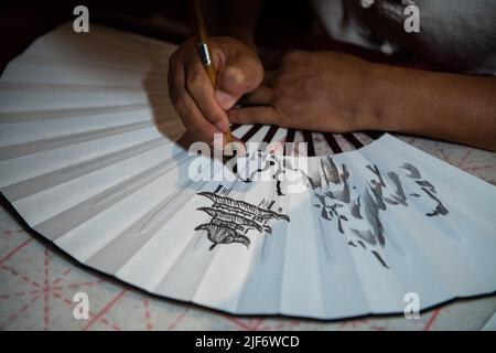 (220630) -- CHANGSHA, June 30, 2022 (Xinhua) -- A painter draws patterns on a fan at the Yueyang tower scenic spot in Yueyang City, central China's Hunan Province, June 23, 2022. Yuezhou fan dates back to the late Ming Dynasty (1368-1644) and early Qing Dynasty (1644-1911). The fan, having a history of about 400 years, is one of the most famous fans in China for its delicacy. Using fine bamboo as its framework, ox horn as the nail, and Xuan paper as the cover, the fan goes through 72 complicated procedures before done, while the making of framework requires most exquisite and skillful work. Th Stock Photo