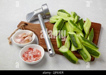 Sliced fresh cucumbers on a wooden cutting board, kitchen hammer, garlic and pink salt on a light concrete table Stock Photo